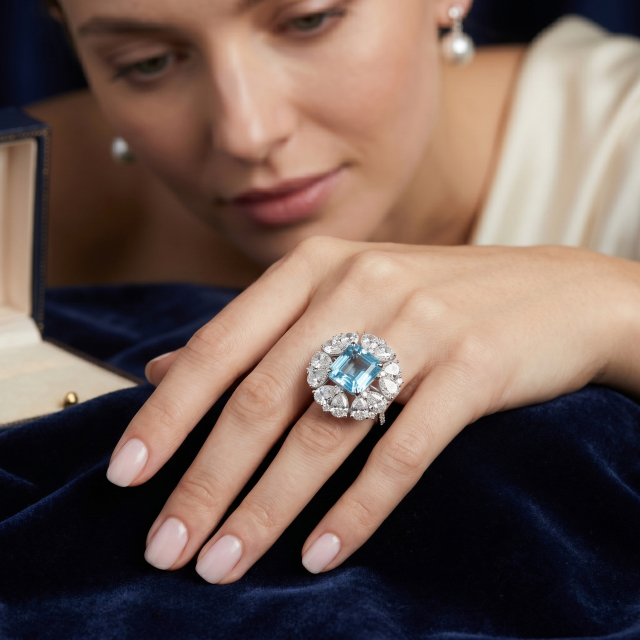 A female model admiring a stunning 14K white gold engagement ring featuring a massive Asscher cut natural London Blue Topaz surrounded by a floral halo of pear-shaped moissanites.