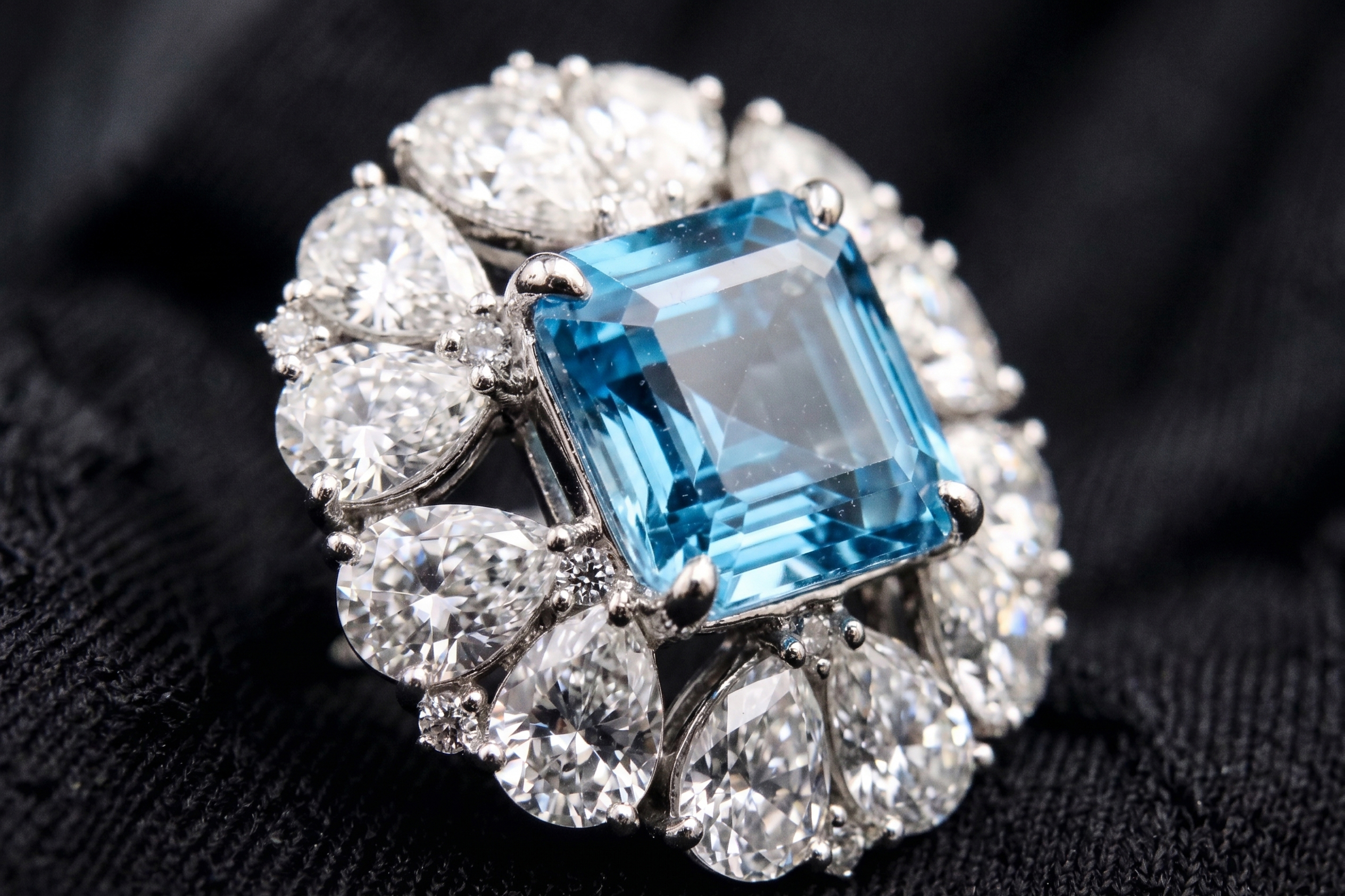 Extreme macro photography on black fabric detailing the step facets of an Asscher cut natural blue topaz and the VVS excellent cut of the 12 surrounding pear-shaped moissanite accent stones.