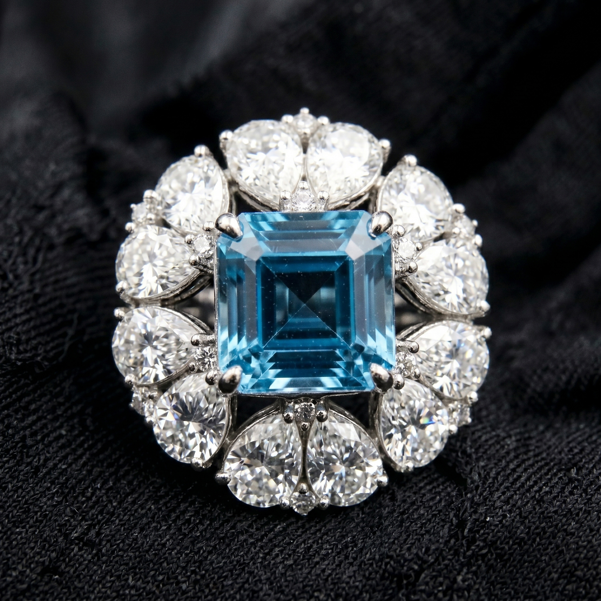 Top-down flat lay view on black texture emphasizing the geometric perfection of the Asscher cut blue topaz center and the brilliant crushed-ice fire of the pear moissanite halo.