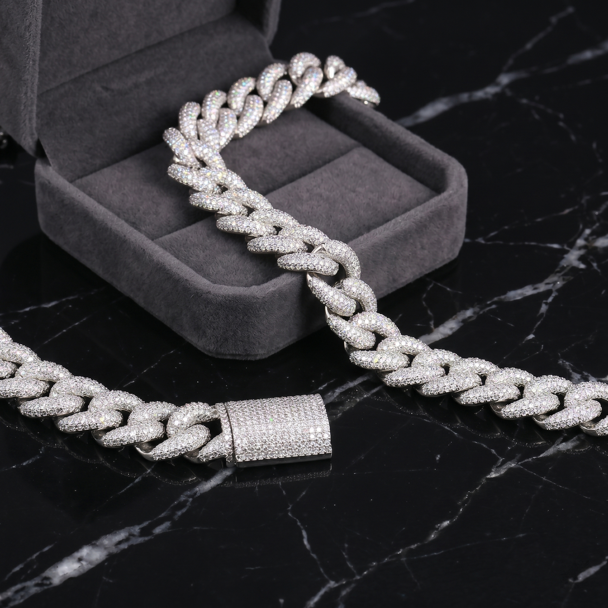 A premium 16mm iced-out moissanite Cuban link chain crafted in S925 sterling silver, draped elegantly from a grey velvet jewelry box onto a black marble surface, highlighting its brilliant diamond-like fire and secure box clasp.