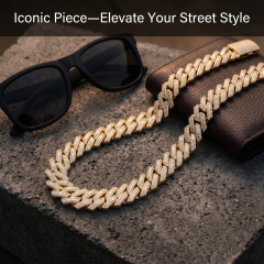 A 16mm yellow gold plated Moissanite Cuban chain presented on a textured concrete base next to luxury sunglasses and a leather wallet.