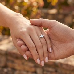 A couple holding hands outdoors, showcasing an elegant 18K gold three-stone engagement ring featuring a pear-cut lab-grown diamond.