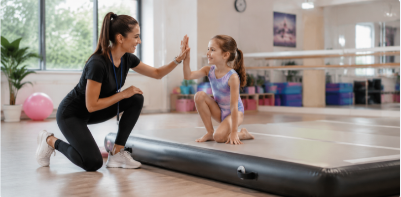 A supportive gymnastics coach encouraging a young child on a safe, cushioned inflatable tumbling mat.