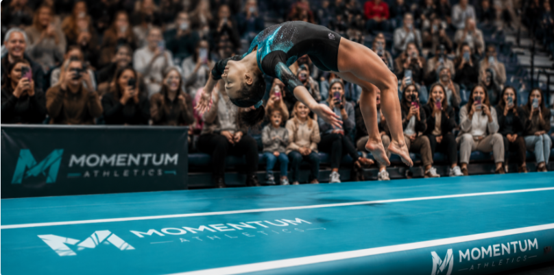 An athlete tumbling over a custom-branded air track mat while spectators film the action on smartphones.