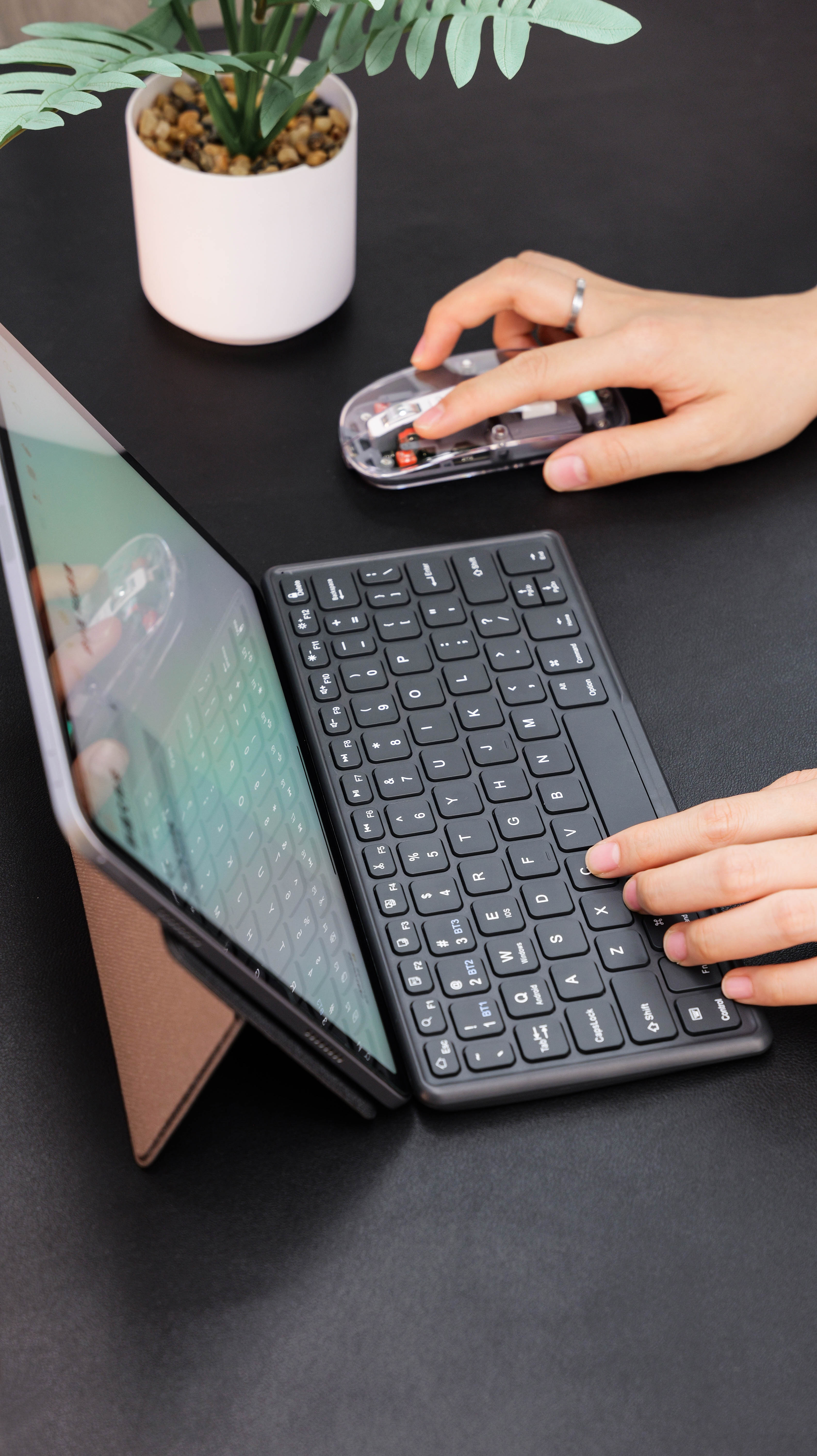 Hands using a compact keyboard and transparent mouse beside a tablet on a desk