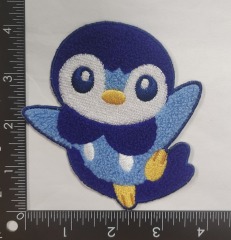 A chenille + embroidery badge featuring a cartoon penguin. The penguin has a white belly, dark blue head, light blue wings, yellow feet and beak, with large round eyes that have blue pupils. The badge has a rounded shape, and a ruler in the background indicates it’s approximately 3.5 inches tall. Chenille gives the penguin’s body a plush, textured look, while embroidery adds details like the beak, feet, and eye outlines.