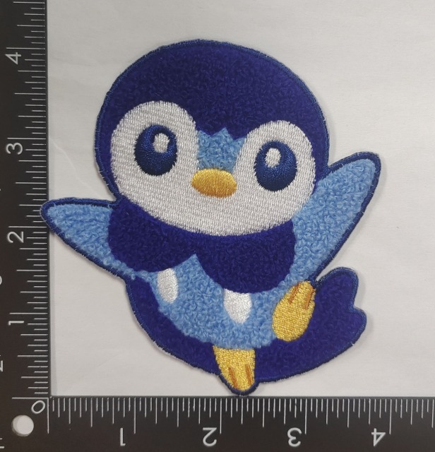 A chenille + embroidery badge featuring a cartoon penguin. The penguin has a white belly, dark blue head, light blue wings, yellow feet and beak, with large round eyes that have blue pupils. The badge has a rounded shape, and a ruler in the background indicates it’s approximately 3.5 inches tall. Chenille gives the penguin’s body a plush, textured look, while embroidery adds details like the beak, feet, and eye outlines.