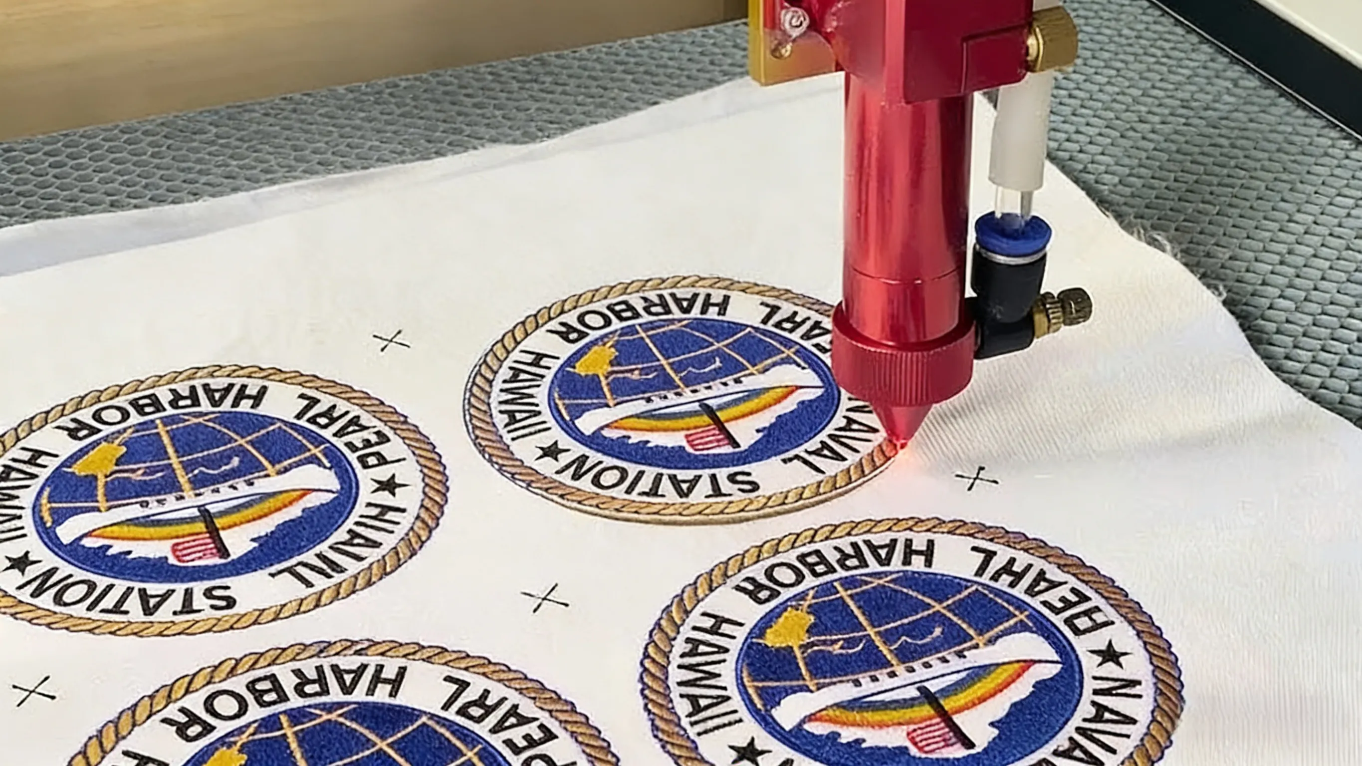 Laser cutting embroidered patches featuring the Pearl Harbor Naval Station Hawaii seal. Precision red laser head trims edges of multi-colored, high-density embroidery on white backing fabric. Ideal for military, nautical, or commemorative apparel.