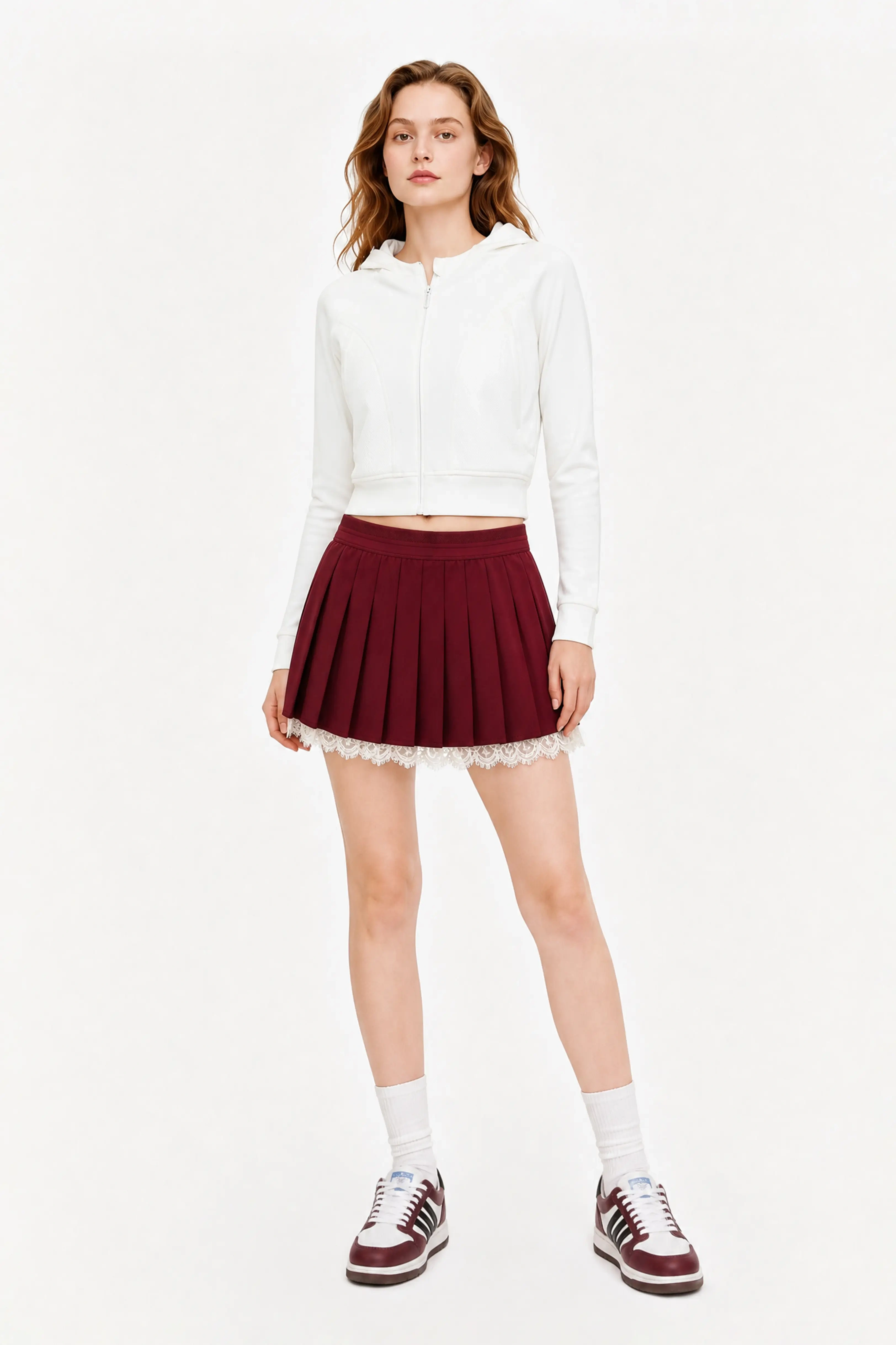 Full body shot of a woman wearing a high waisted burgundy tennis skirt paired with a white hoodie and sneakers.