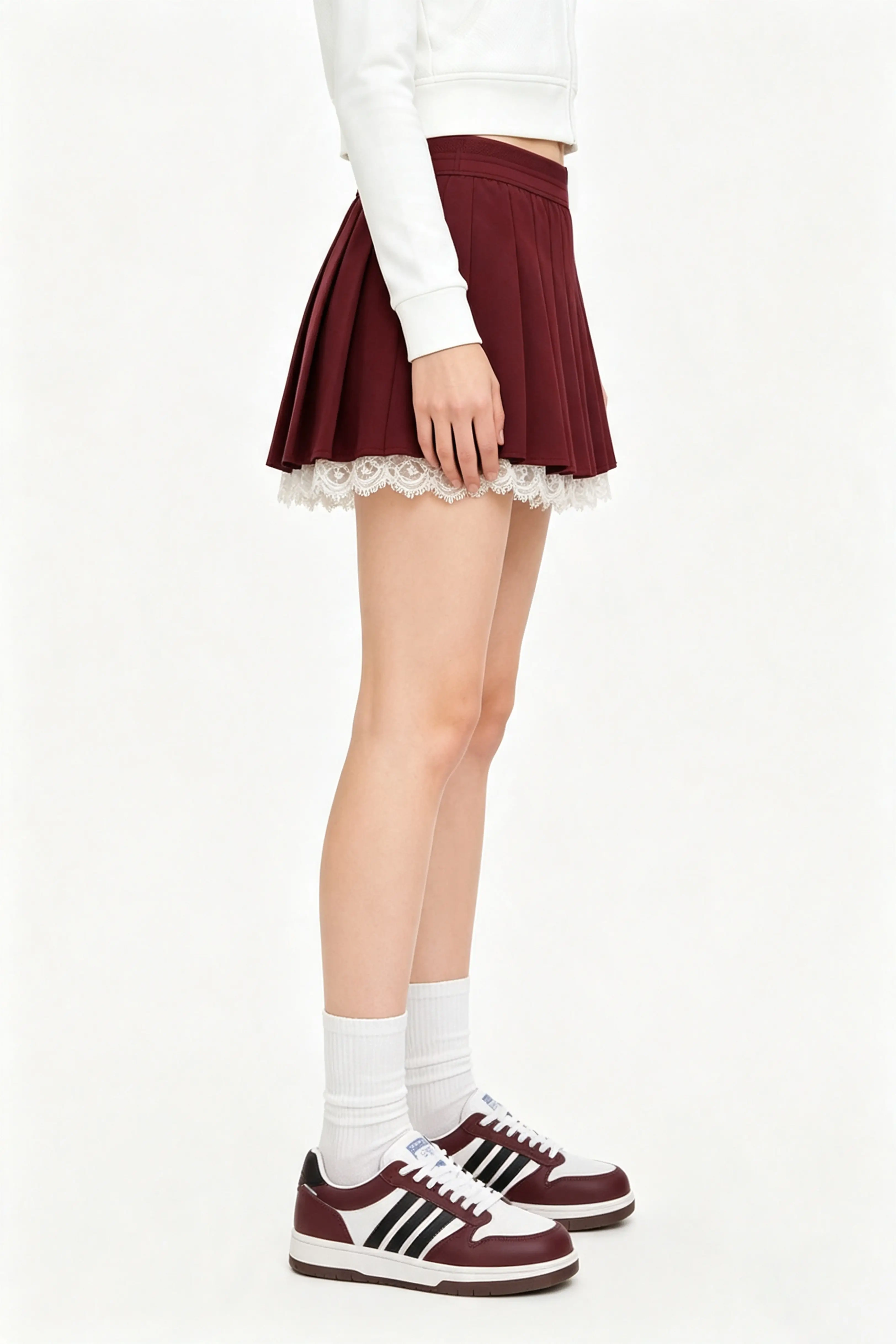 Side view of a model showcasing the fit of a high waisted burgundy pleated tennis skirt with white lace hem.