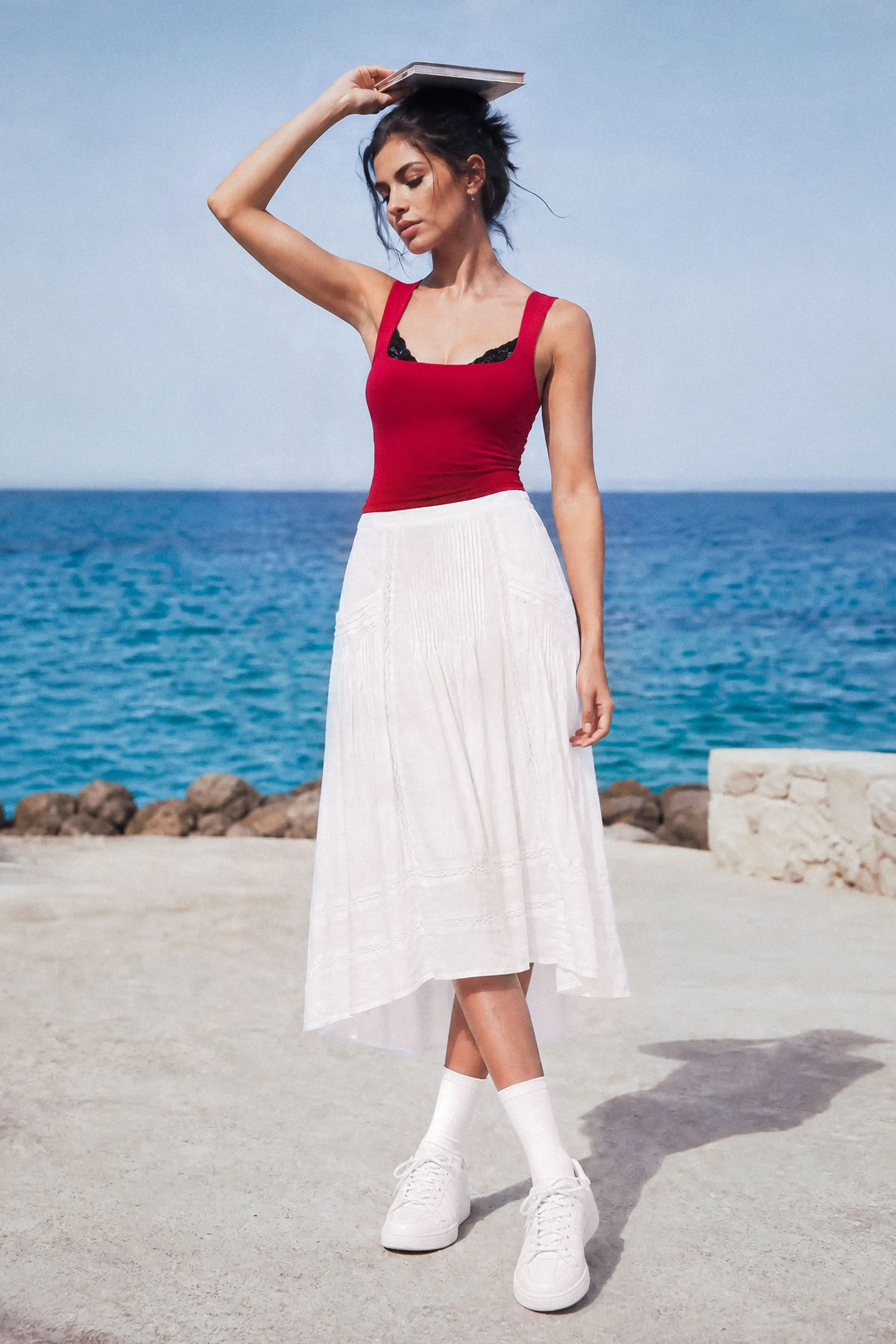 A woman posing by the sea holding a book, wearing a white pleated midi skirt with a red tank top and white sneakers.