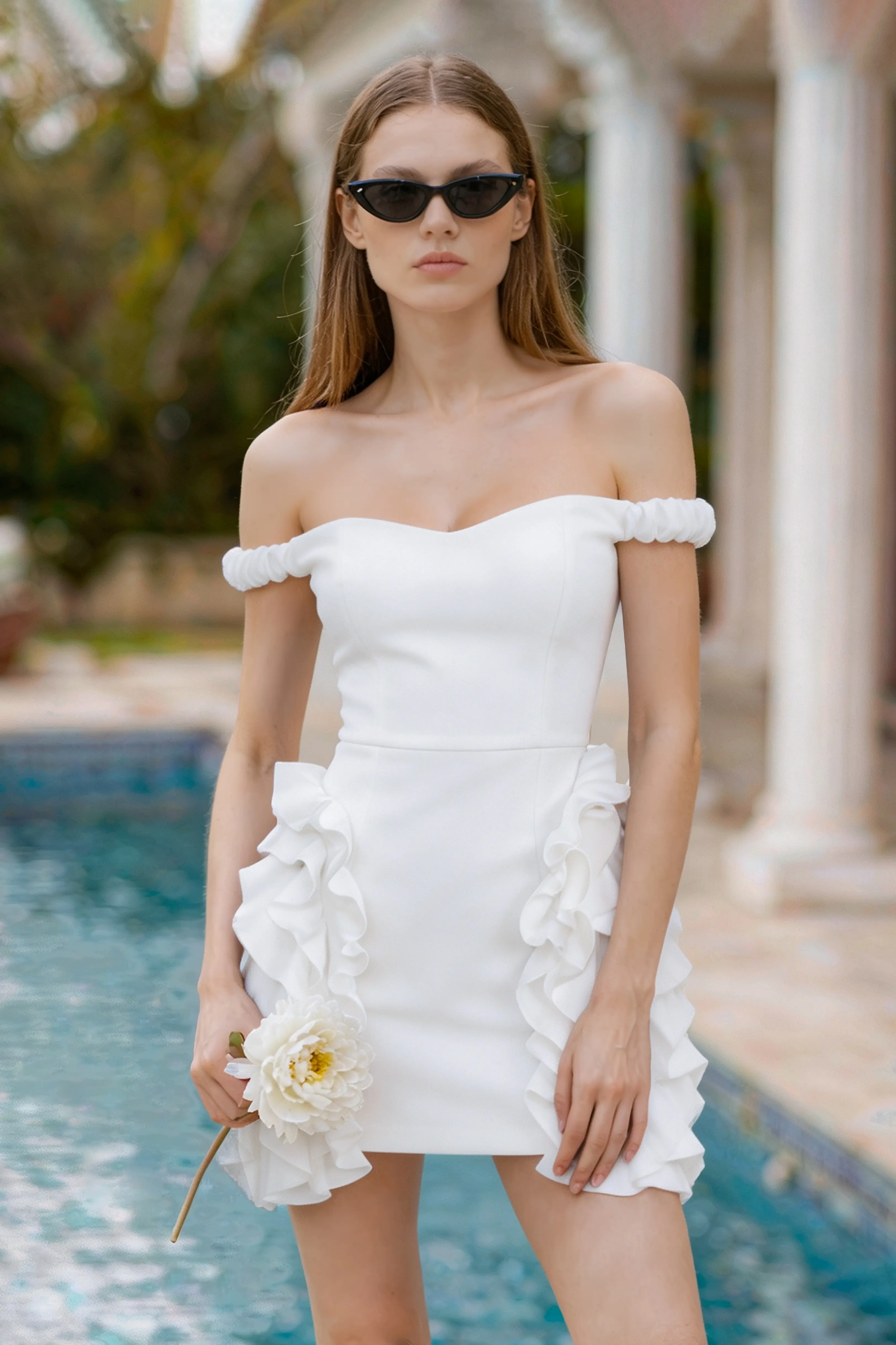 A woman posing by a pool holding a white flower, wearing an off-the-shoulder white ruffle mini dress with sunglasses.