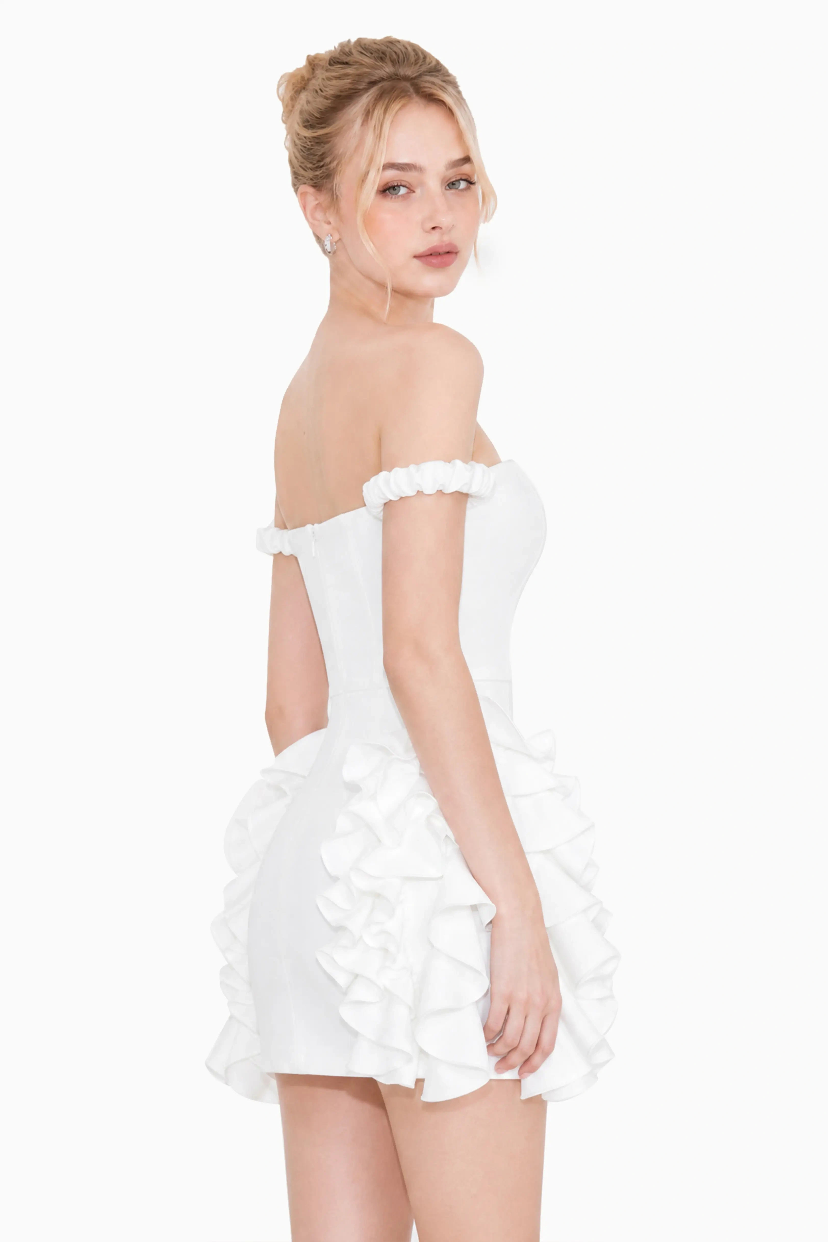 Side profile view of a woman wearing a fitted white ruffle mini dress with off-the-shoulder straps and tiered side ruffles.