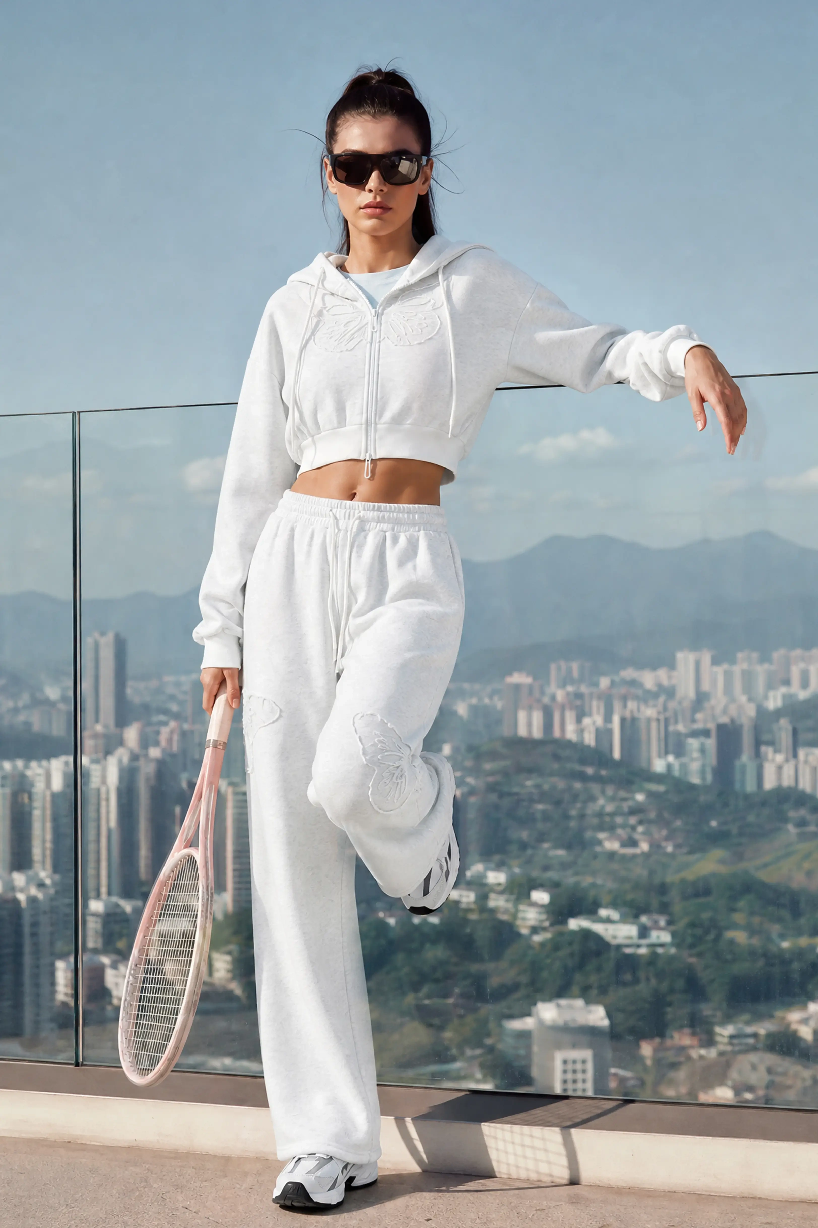 A woman holding a tennis racket on a rooftop, wearing a grey cropped zip-up hoodie and high-waisted heather grey butterfly sweatpants.