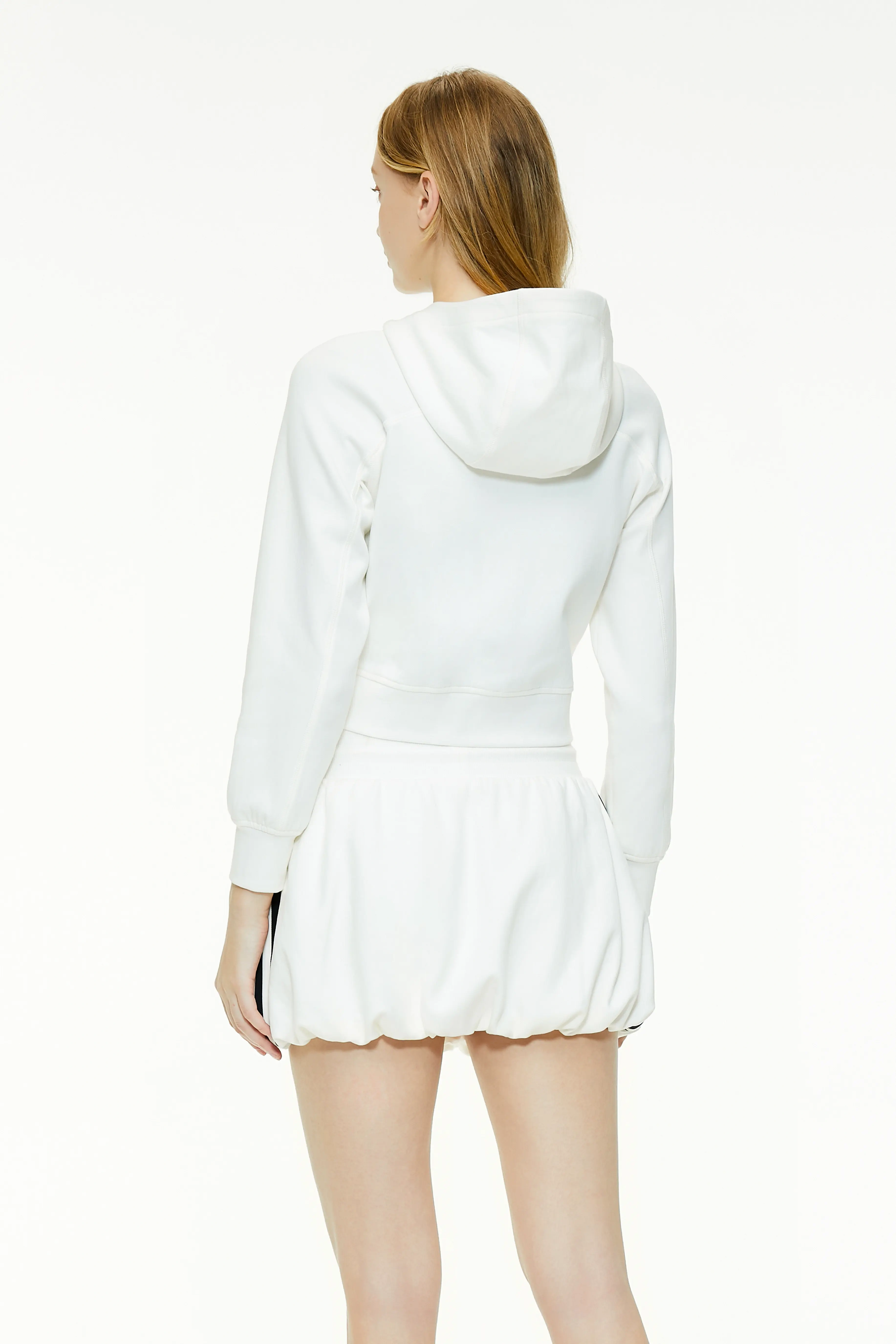 Back view of a white cropped zip-up hoodie showcasing the relaxed hood, fitted shoulders, and bubble skirt silhouette.