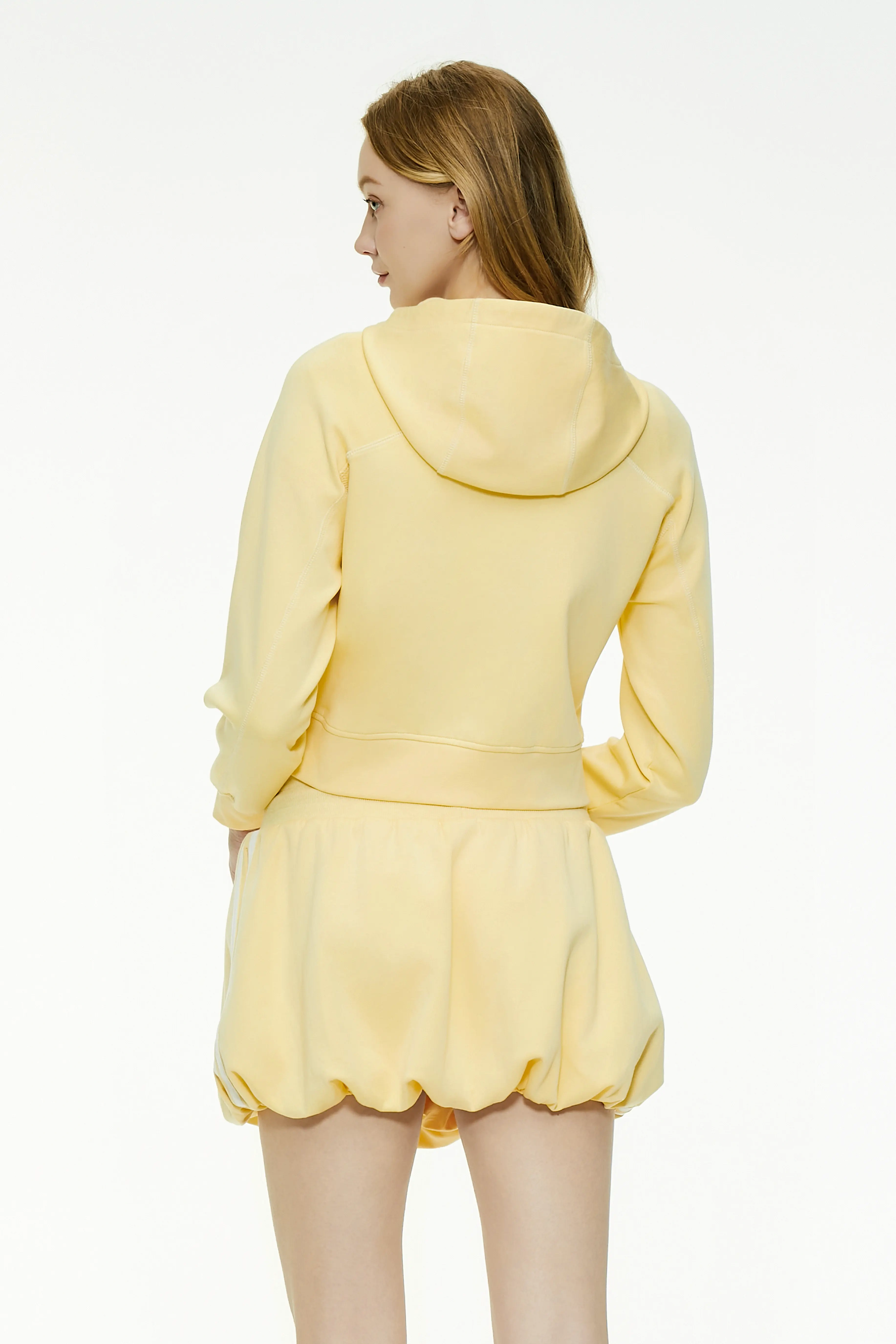 Back view of a pale yellow cropped zip-up hoodie featuring a hood and clean rear silhouette paired with a bubble skirt.