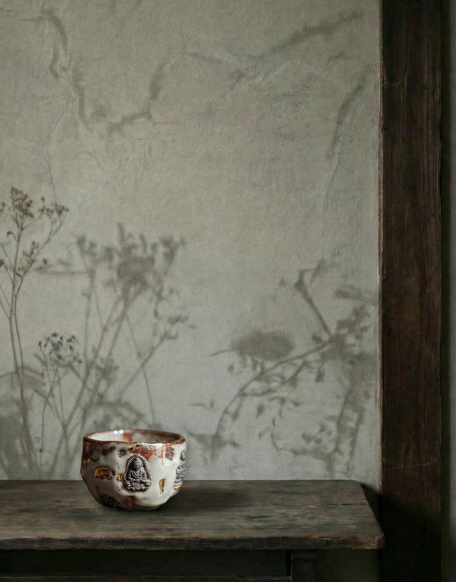 A close-up of a handmade ceramic tea cup with a textured surface and an embossed Buddha figure. The cup sits on a rustic wooden table against a minimalist grey-green wabi sabi decor wall with delicate plant shadows. To the right, a dark vertical wooden beam provides a structured contrast, creating a peaceful and minimal interior atmosphere.