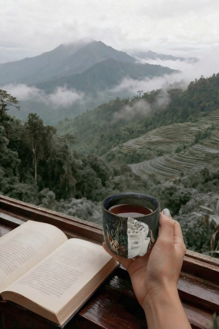 Hand holding a Buddha-adorned teacup by the window, with an open book beside, overlooking mist-shrouded mountains and lush forests&mdash;a tranquil blend of Zen, reading, and nature&rsquo;s serenity.
