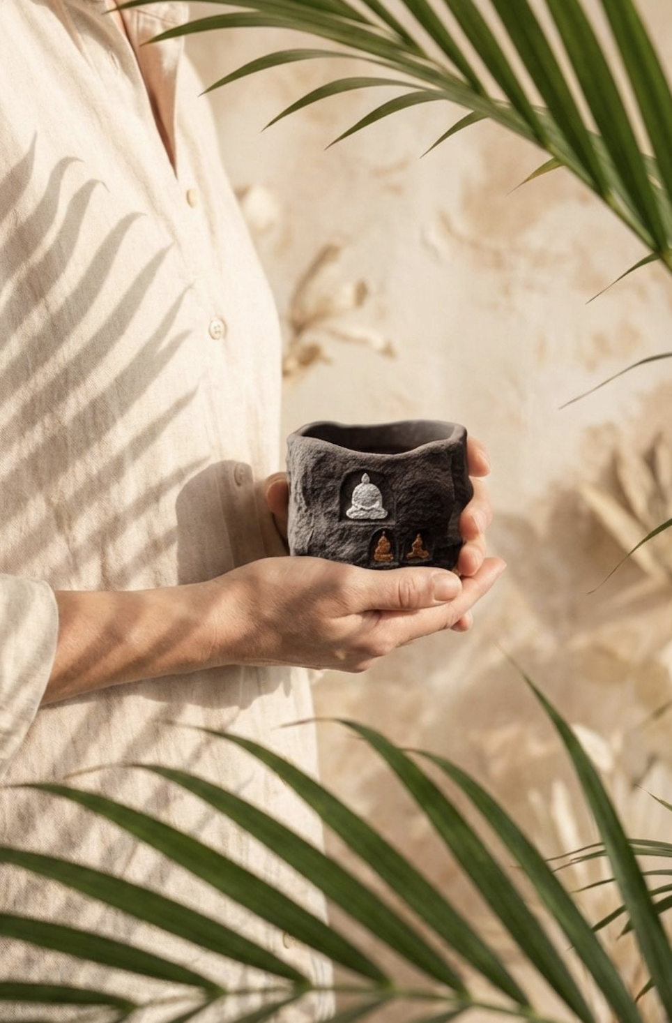 A person in a cream linen shirt holds a handcrafted small batch ceramic vessel featuring ice crackle glaze and a tiny gold Buddha accent, surrounded by dappled sunlight and palm leaves. This luxury self care gift for women doubles as a tactile mindfulness tool, ideal for gifting to moms or anyone craving artisanal, soulful home decor.