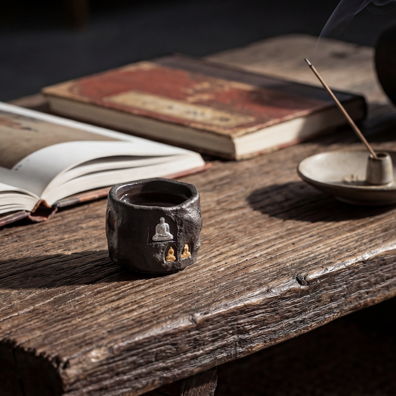 Handcrafted wood-fired ceramics tea cup with embossed Buddha motifs resting on a rustic wooden table beside burning incense and vintage books—perfect example of wabi sabi pottery for a mindful tea ritual that encourages natural patina development over time.