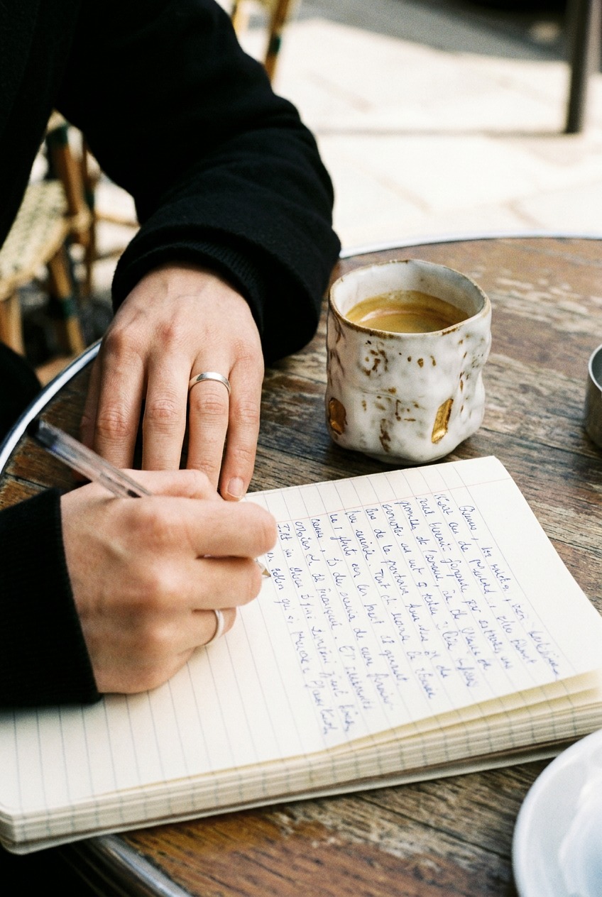 A handmade white ceramic cup with gold accent windows sitting on a round wooden cafe table as a person writes by hand in an open notebook during a morning journaling session.
