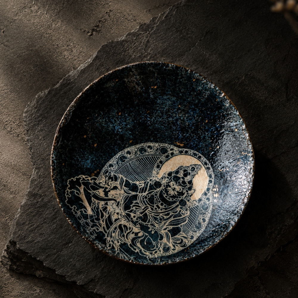 Top-down view of the Midnight Deity ceramic plate on a dark natural stone slab, showcasing the deep navy glaze, subtle gold dusting, and intricately engraved deity relief in tactile wabi-sabi style.