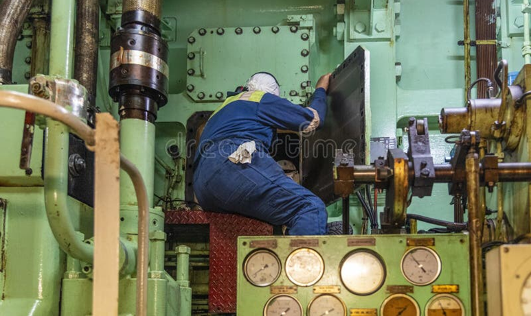 Marine engineer inspecting heavy machinery and mechanical components inside a noisy ship engine room.