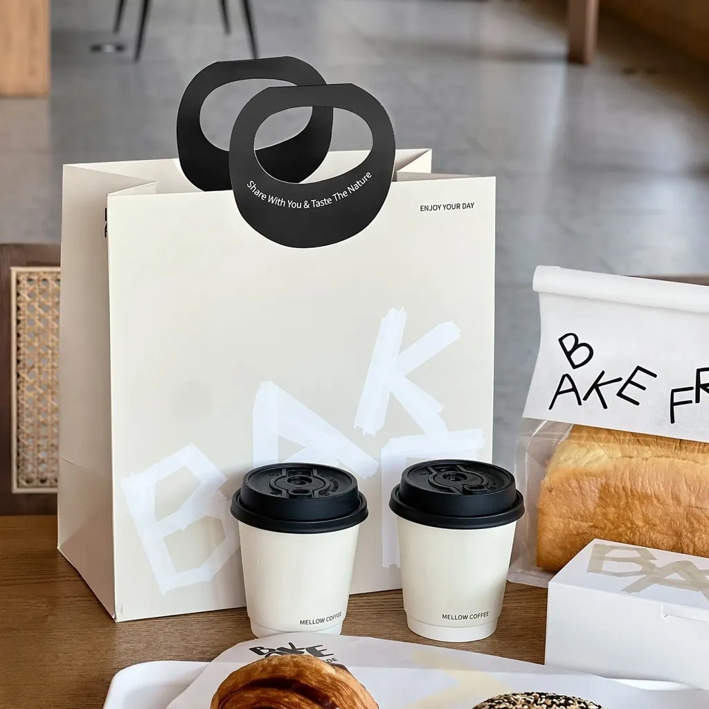 White paper carrier bag with black die-cut handles and printed coffee cups for bakery takeaway