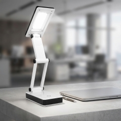 An adjustable white and black folding COB LED desk lamp illuminated on an office desk next to a laptop and pen