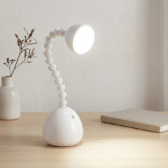 A white LED desk lamp with a unique flexible jointed gooseneck neck illuminating a wooden table next to a book and small vase