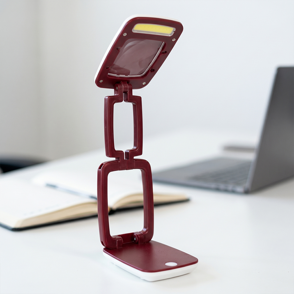 An adjustable burgundy folding LED desk lamp with an integrated magnifying glass lens on a white office desk with a blurred laptop background