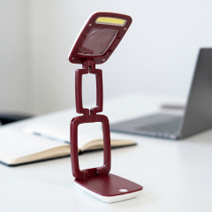 An adjustable burgundy folding LED desk lamp with an integrated magnifying glass lens on a white office desk with a blurred laptop background