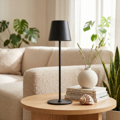 A sleek black metal cordless table lamp on a round wooden side table, next to a stack of books titled Hygge and Minimal Design, a white ceramic vase with grass, and a large seashell in a bright modern living room.