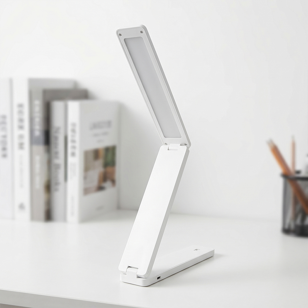 A sleek white folding LED desk lamp in a minimalist design standing on a white table with blurred books and a pencil holder in the background