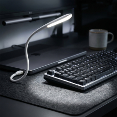 A flexible white USB gooseneck LED lamp illuminating a mechanical keyboard on a desk setup with a laptop and coffee mug in the background