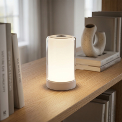 A sleek white cylindrical table lamp illuminating a wooden shelf with books and modern decor in warm white light