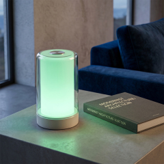A cylindrical LED night lamp glowing soft green on a side table next to a book and a blue sofa