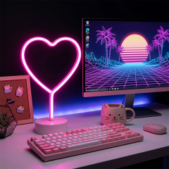 An illuminated pink neon heart table lamp placed on a white gaming desk next to a mechanical keyboard and a computer monitor