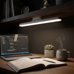 An illuminated dimmable reading light bar mounted under a shelf, lighting up an open book and a laptop screen on a cozy dark wooden desk