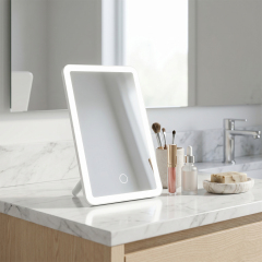 A square white LED makeup vanity mirror standing on a marble bathroom countertop next to cosmetics and a faucet