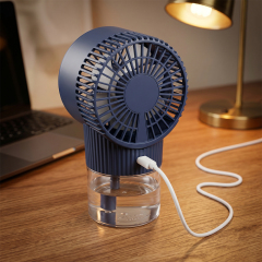 Rear angled view of a dark blue desktop misting fan connected to a white USB charging cable on a wooden desk next to a table lamp