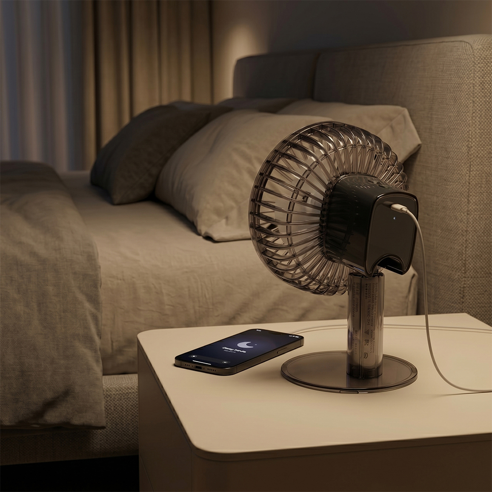 A smoked-black bedside table fan connected to a charging cable on a nightstand in a dimly lit bedroom setting