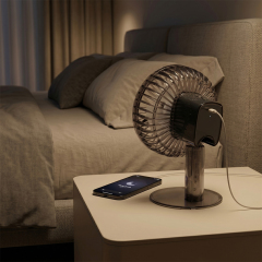 A smoked-black bedside table fan connected to a charging cable on a nightstand in a dimly lit bedroom setting