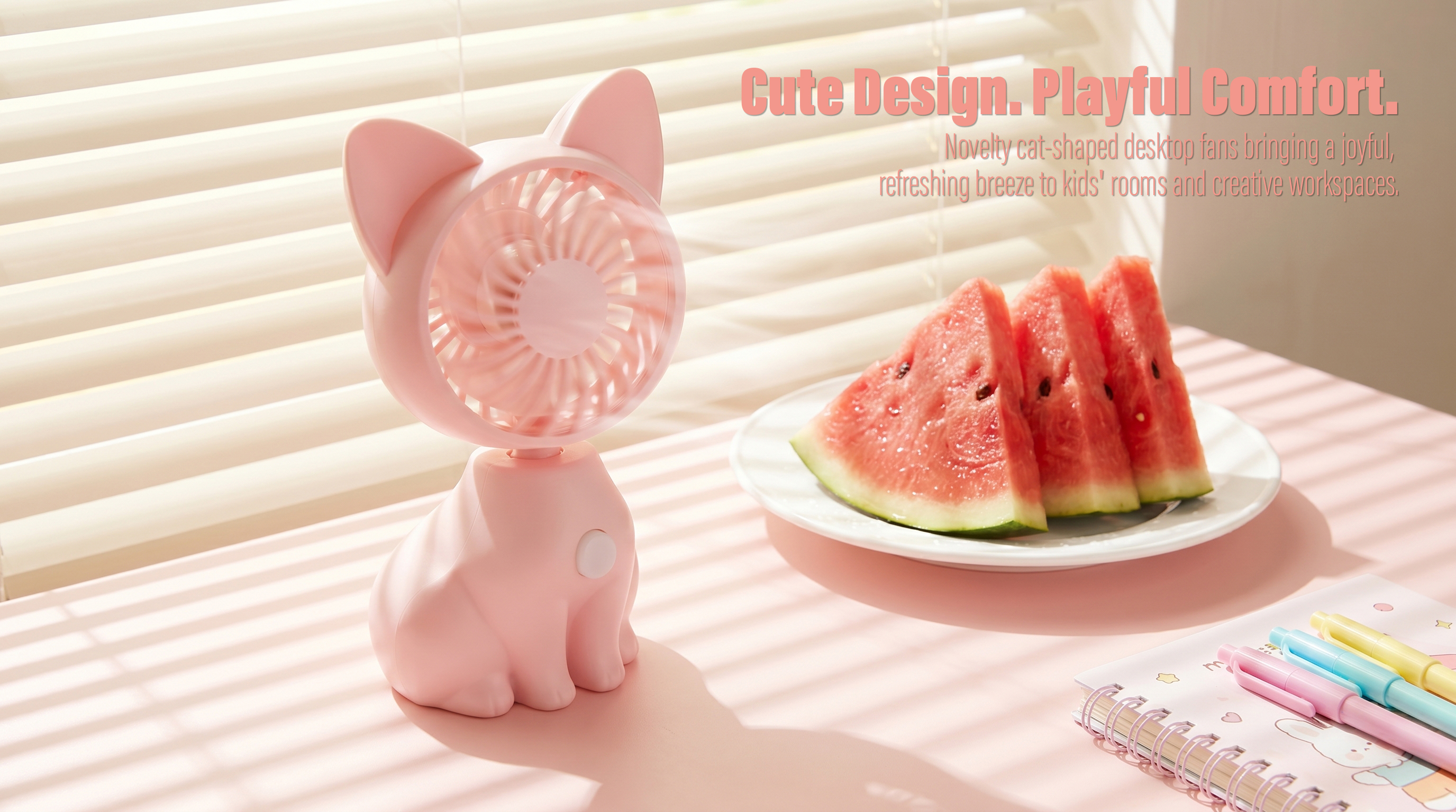 A pink cat-shaped desktop mini fan operating on a pink table under soft window blind shadows next to a white plate of sliced watermelon and a notebook with colorful pens.