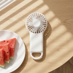 A white handheld mini fan resting on a light-colored wooden table under soft window blind shadows next to a white plate of sliced watermelon.