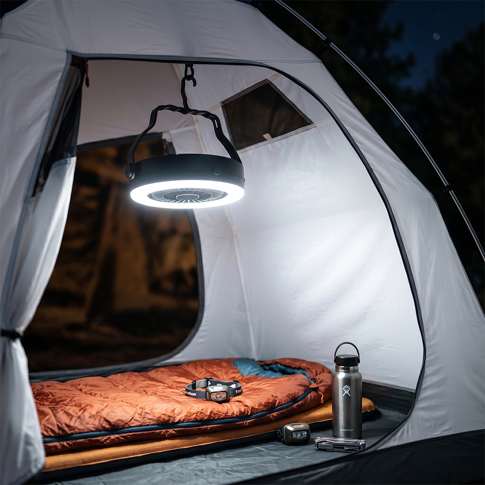 A 2-in-1 camping fan hanging from the ceiling of a lit tent at night, illuminating a sleeping bag and a thermos bottle setup.
