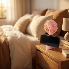 A pink desktop misting fan with a smartphone resting in its base cradle, placed on a wooden nightstand next to a bed with soft pillows in a cozy bedroom.