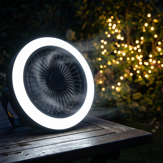 The circular outdoor fan operating on a wooden table at night with its bright white LED ring lit up against a blurred background of warm fairy lights.