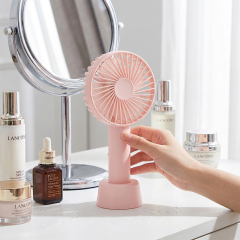 A person's hand placing a pink mini fan onto its base on a white vanity table next to a silver makeup mirror and skincare bottles.