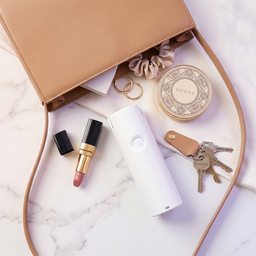 Flat lay view of a white pocket-sized telescopic mini fan sitting next to a leather handbag, lipstick, and keys on a marble surface.